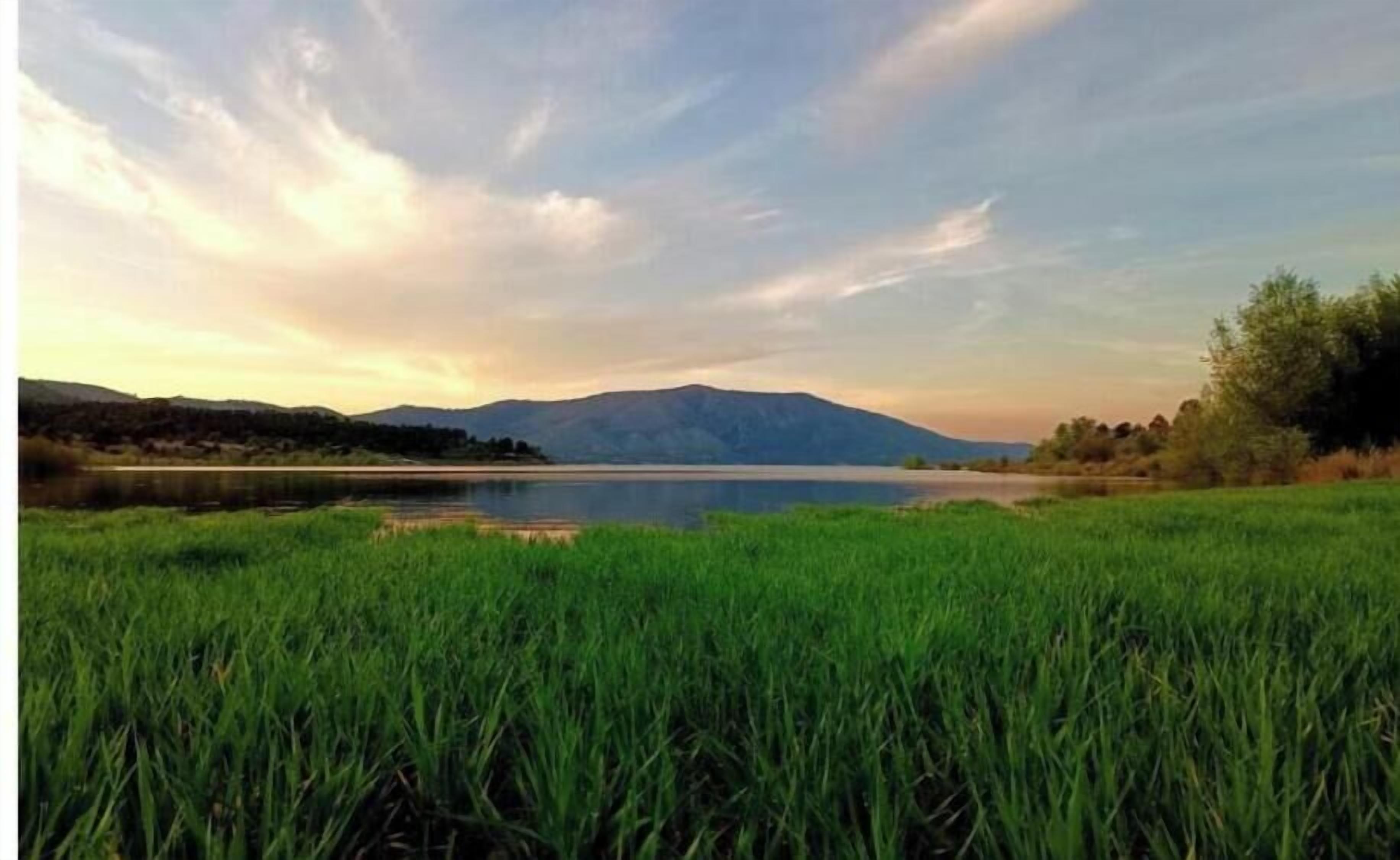 Un paisaje sereno con un lago, un primer plano de hierba y montañas distantes bajo un cielo parcialmente nublado.
