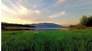Un paisaje sereno con un lago, un primer plano de hierba y montañas distantes bajo un cielo parcialmente nublado.