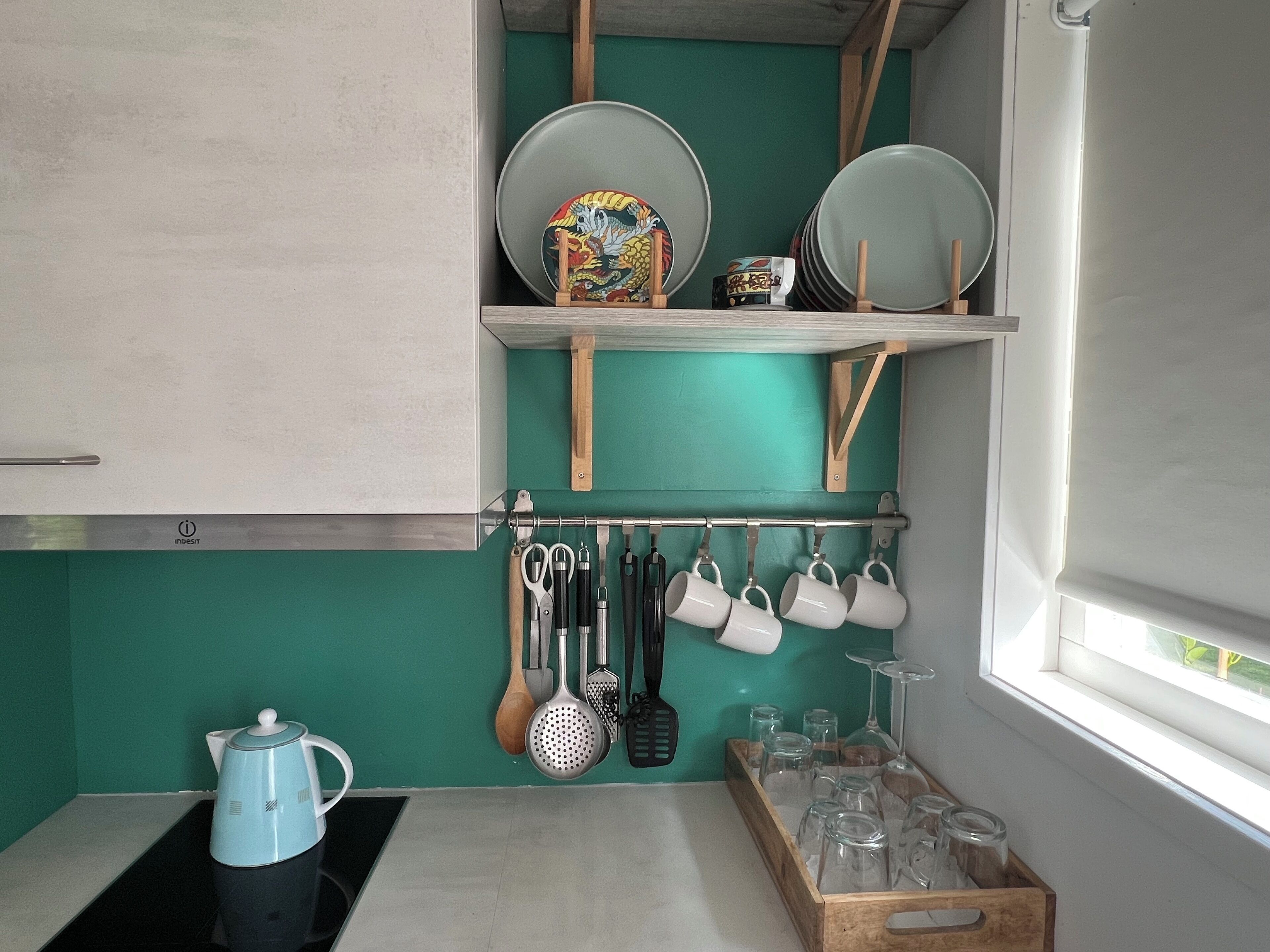 A kitchen with a teal backsplash, a coffee maker, hanging utensils, and a rack of plates and cups.