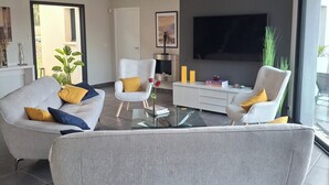 A modern living room with a grey sofa, a white armchair, a glass coffee table, and a flat-screen TV mounted on the wall. - Superb contemporary villa portes Perigord 160m2, air-conditioned, swimming pool, lake 2500m away. (Chasteaux)