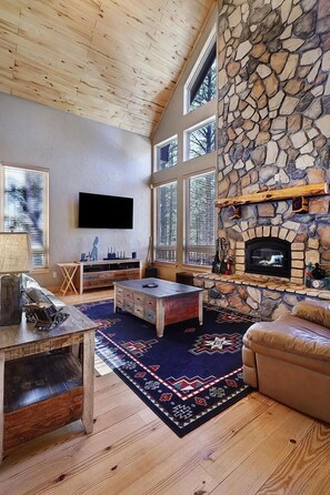 A cozy living room with a stone fireplace, wooden ceiling, and large windows. - Large kitchen, pool table, families, 5 mil to biking (Angel Fire)