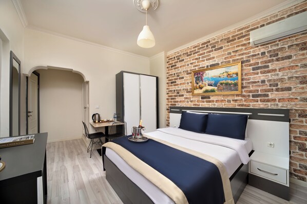 A hotel room with a brick wall, a bed with a headboard, a desk with a chair, a mirror, and a painting.