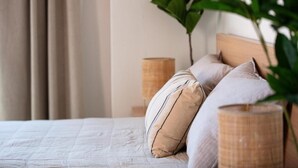 A bed with a beige quilt, two pillows, and a wooden headboard. There is a plant in the foreground and a lamp on the nightstand.