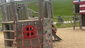Eine Holzspielkonstruktion mit Kletternetzen und einer Rutsche, vor einem sandigen Strand und einem grünen Rasen.