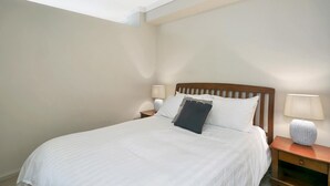 Una cama bien tendida con sábanas blancas y un almohadón oscuro, un cabecero de madera y dos lámparas de noche sobre mesitas de madera.