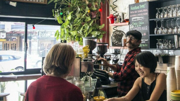Interior de una cafetería con un barista preparando bebidas, un cartel de menú y vista a la calle a través de una ventana.