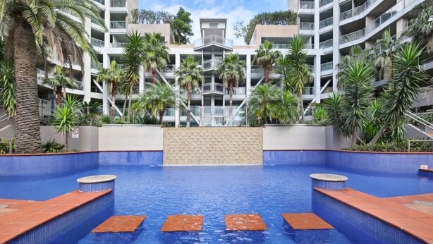Una piscina con piso azul de baldosas, rodeada de palmeras y un edificio de varios pisos con balcones.
