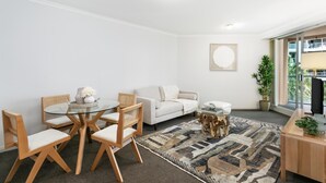 Una sala de estar moderna con una mesa de comedor de vidrio, sillas de madera, un sofá blanco, un tapete estampado y un ventanal con vistas a edificios.
