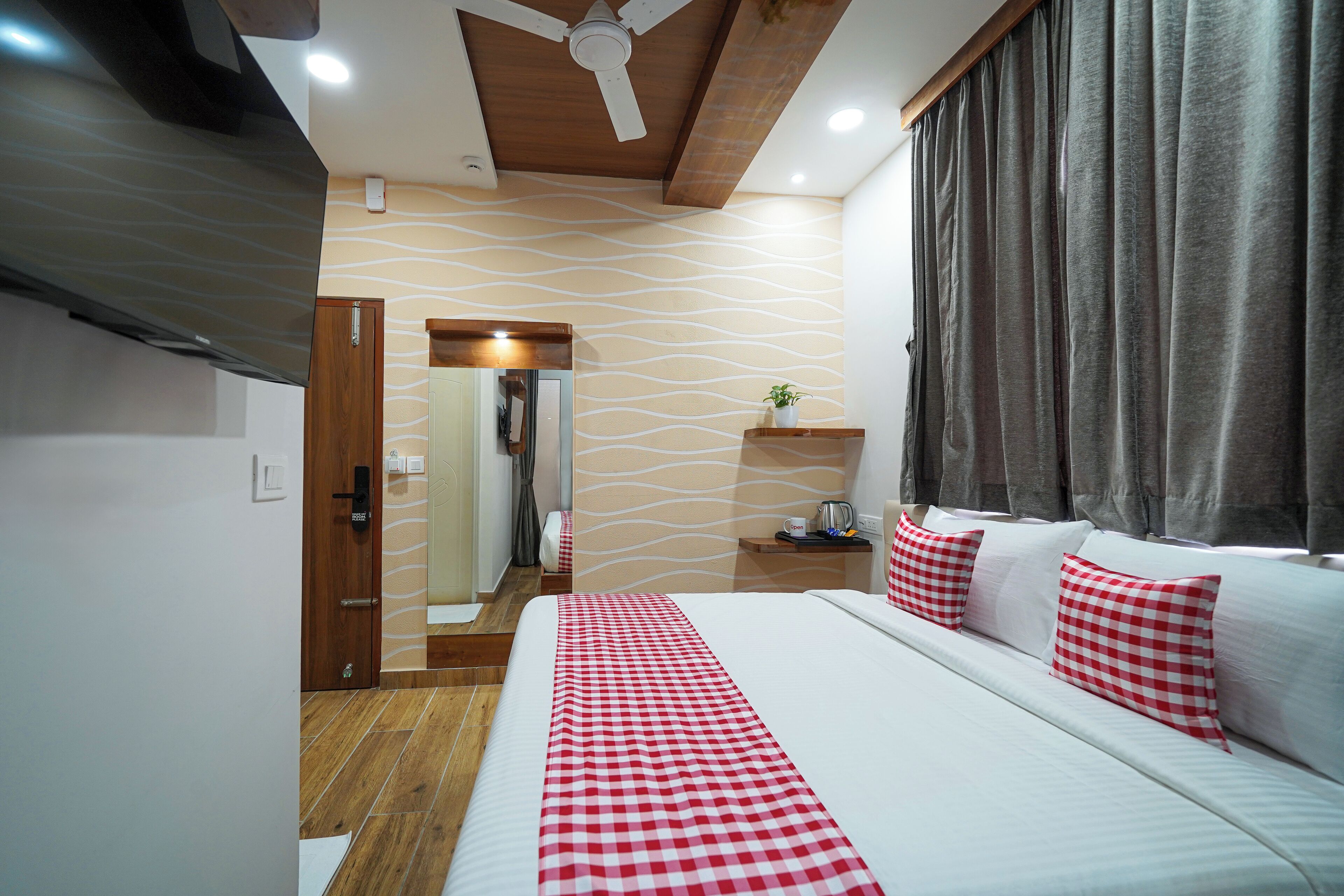 A hotel room with a bed, a wooden ceiling, a wall with a wavy pattern, a bedside table with a plant, and a window with curtains.