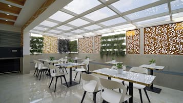 A modern indoor dining area with a glass ceiling, white and black chairs, and tables with potted plants.