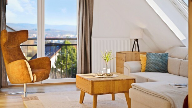 A modern living room with a high ceiling, a large window with a view of the city, a brown leather chair, a wooden coffee table, and a beige sofa with colorful cushions. - Comfort kisses nature Skyline vacation apartment Goldbach (Goldbach)