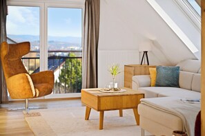 A modern living room with a high ceiling, a large window with a view of the city, a brown leather chair, a wooden coffee table, and a beige sofa with colorful cushions. - Comfort kisses nature Skyline vacation apartment Goldbach (Goldbach)