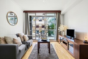 A modern living room with a sofa, a coffee table, a TV on a wooden cabinet, and a large window with a view of buildings. - Sydney - KS004 - Kent St - CBD crib | Skyline views meet pool & gym living (Sydney)