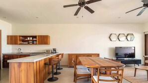 A modern kitchen with a central island, bar stools, a dining table with chairs, a ceiling fan, and a television in a living area.