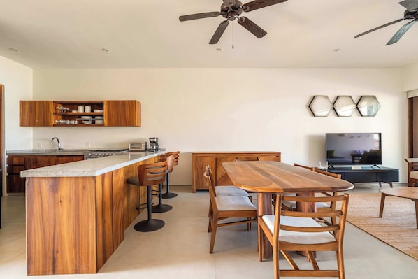 A modern kitchen with a central island, bar stools, a dining table with chairs, a ceiling fan, and a television in a living area.