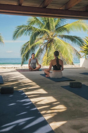 Two individuals seated on mats outdoors, with a palm tree and ocean view.