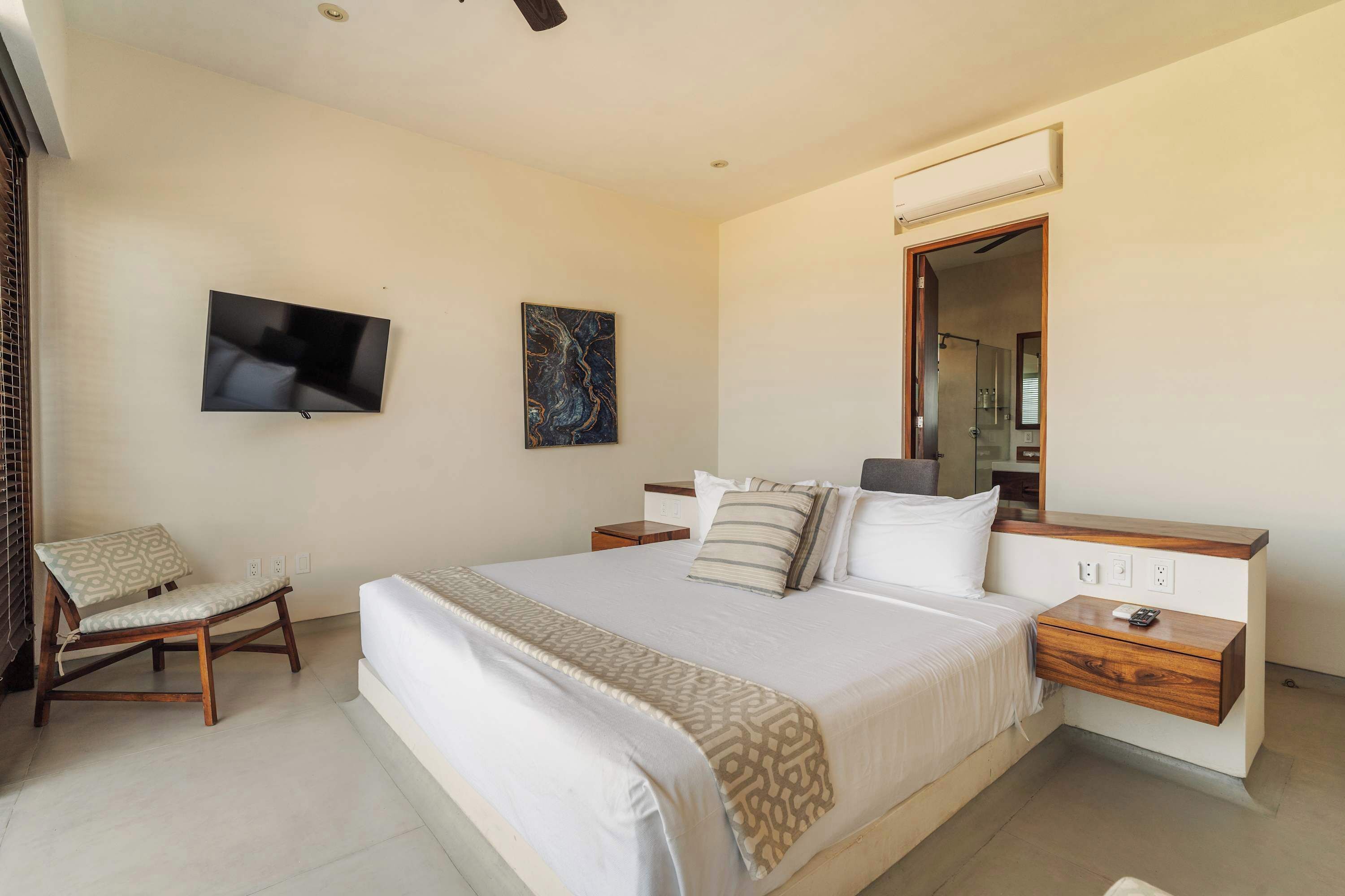 A hotel room with a bed, a TV, a painting, a chair, and a wooden nightstand.