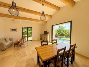 A spacious room with a wooden dining table, chairs, a sofa, and a large window overlooking a swimming pool.