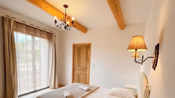 Une chambre à coucher avec un lit, une table de chevet, une lampe, un lustre et une fenêtre avec des rideaux.