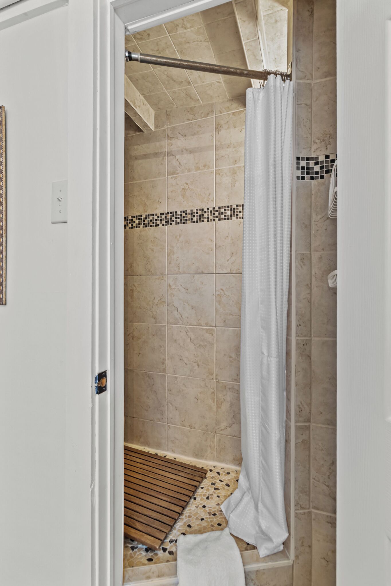 A shower area with beige tiles, a white shower curtain, and a wooden bench.