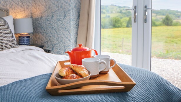 Un lit recouvert d’une couverture bleue, un plateau en bois avec des croissants, une théière rouge et des tasses blanches.