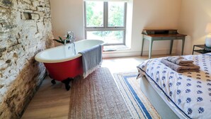 Une chambre avec une grande fenêtre, une baignoire rouge, un bureau en bois et un lit recouvert d’une couvre-lit à motifs.