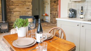 Un coin repas avec une table en bois dressée pour deux personnes, une bouteille de vin et une plante en pot. On aperçoit un mur de pierre et une cheminée à l’arrière-plan.
