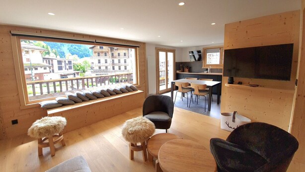 Ein modernes Wohnzimmer mit großem Fenster, Holzmöbeln, einem Flachbildfernseher und einer Küche im Hintergrund.