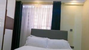 A neatly made bed with a white comforter and pillows, a headboard, and a window with sheer curtains and a dark drape. - Shirpelle Suites Kiamunyi (Nakuru)