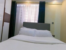 Una cama bien hecha con una colcha blanca y almohadas, un cabecero y una ventana con cortinas translúcidas y un cortinaje oscuro.