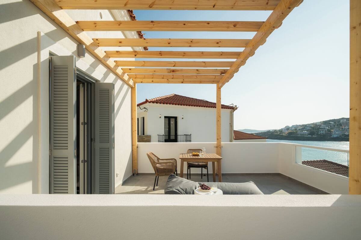 A balcony with a wooden pergola, a table with chairs, a sofa, and a view of a building and coastline.