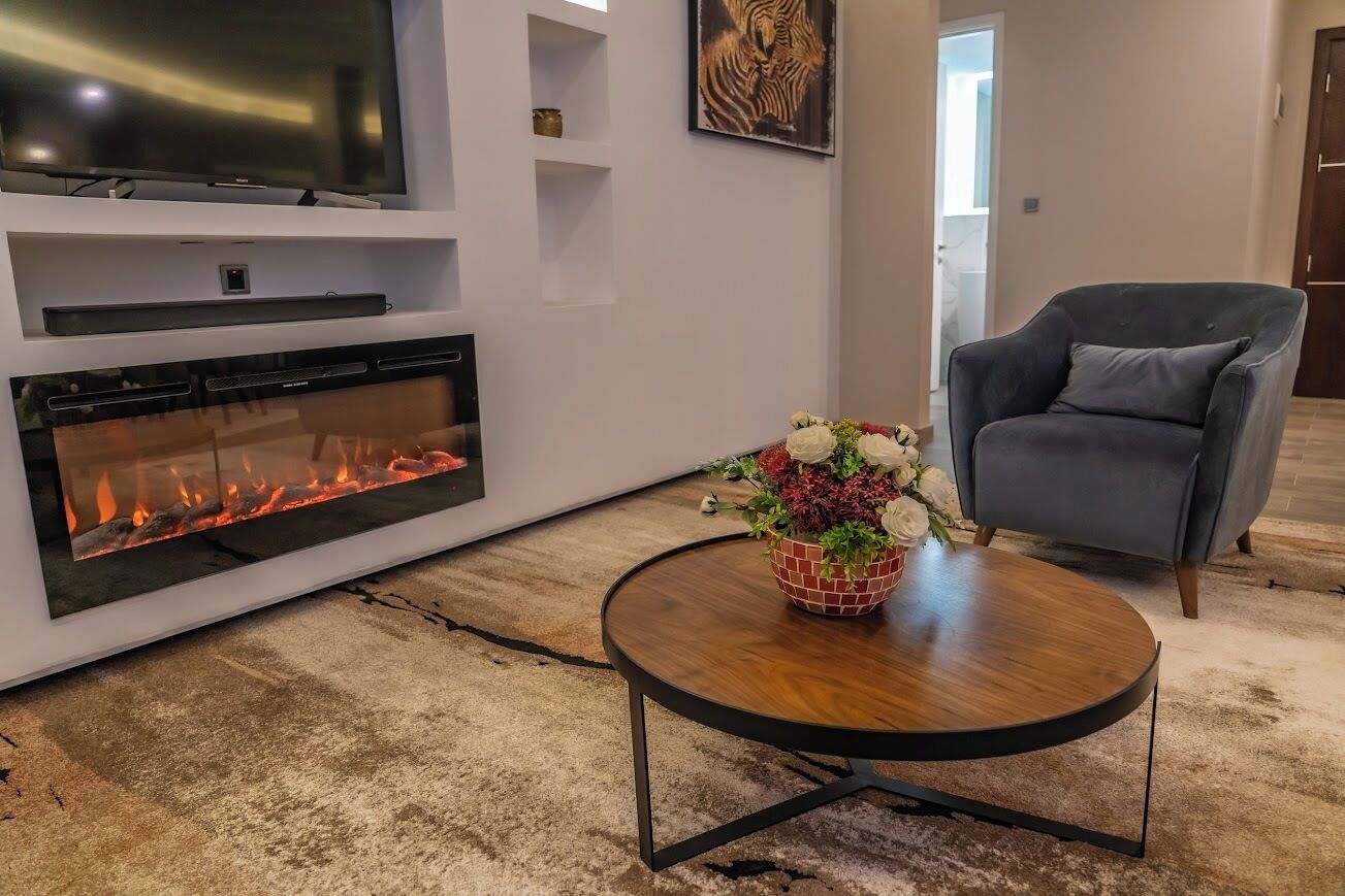 A modern living room with a fireplace, a flat-screen TV, a grey armchair, and a round wooden coffee table with a floral centerpiece.