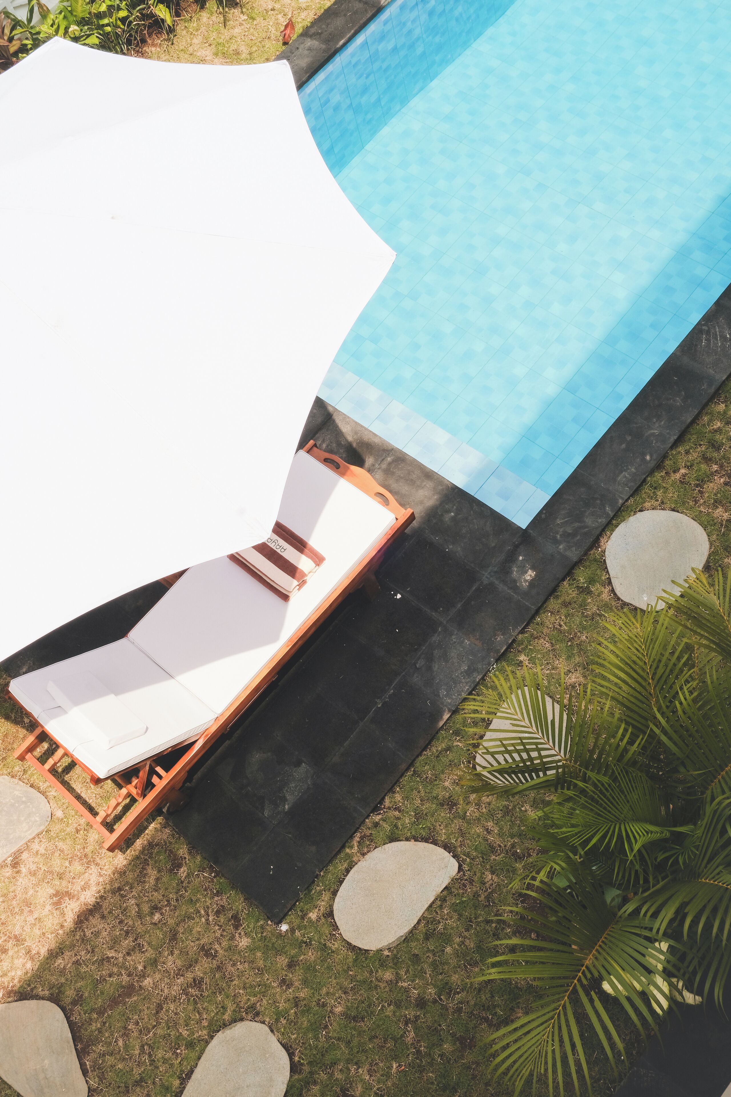 A poolside area with a lounge chair and umbrella, surrounded by a stone border and greenery.