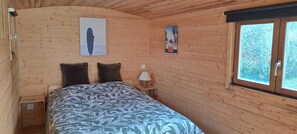Een slaapkamer in een houten chalet, met een bed, twee kussens, een nachtkastje met een lamp en twee aan de muur hangende foto's.