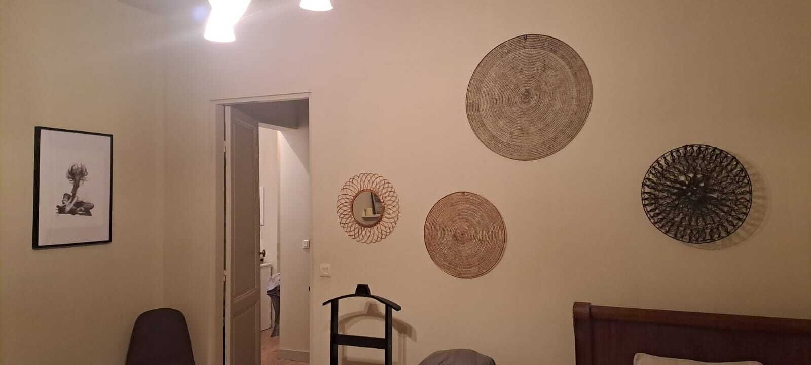 A room with a bed, a chair, and a door leading to another room. There are wall decorations including a framed picture and three circular wall hangings.
