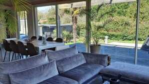 A modern outdoor seating area with a grey sofa and dining table, surrounded by large windows offering a view of a swimming pool and greenery. - House with heated pool Soulac 4bed near Ocean (Soulac-sur-Mer)