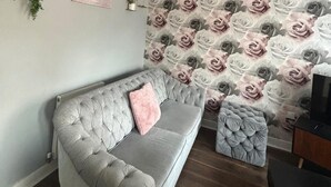 A grey tufted sofa with a pink pillow against a wall with a floral wallpaper pattern. - Trowbridge House, Long Stay Disc, Parking, Wifi (Swindon)
