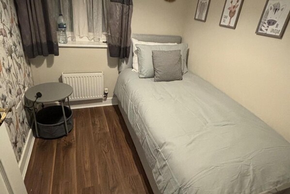 A small, single-bed room with a round table, a radiator, and a window with curtains.