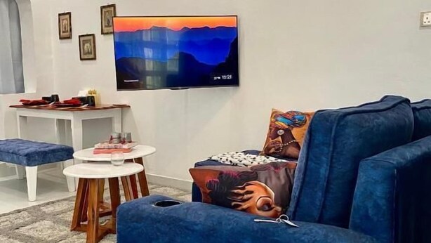 Un salon avec un canapé bleu, un téléviseur fixé au mur, une petite table ronde et un mur blanc orné de tableaux encadrés.