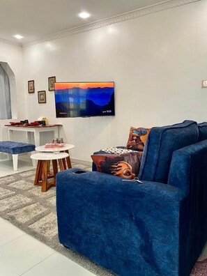 Una sala de estar con un sofá azul, un televisor montado en la pared, una pequeña mesa redonda y una pared blanca adornada con marcos.