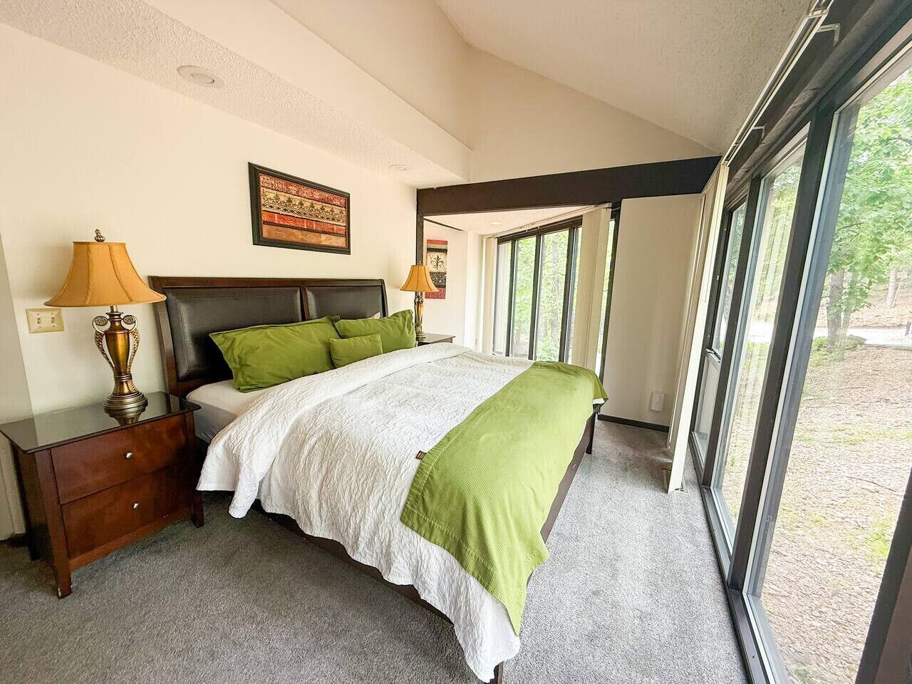 A bedroom with a bed, nightstands, lamps, a framed picture, and large windows.