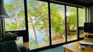 Una habitación con amplios ventanales que dan a un lago y árboles, una mesa de madera y un sofá verde.