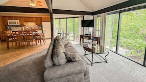 A spacious living room with a large window, a sofa, a TV, and a dining area with wooden furniture.