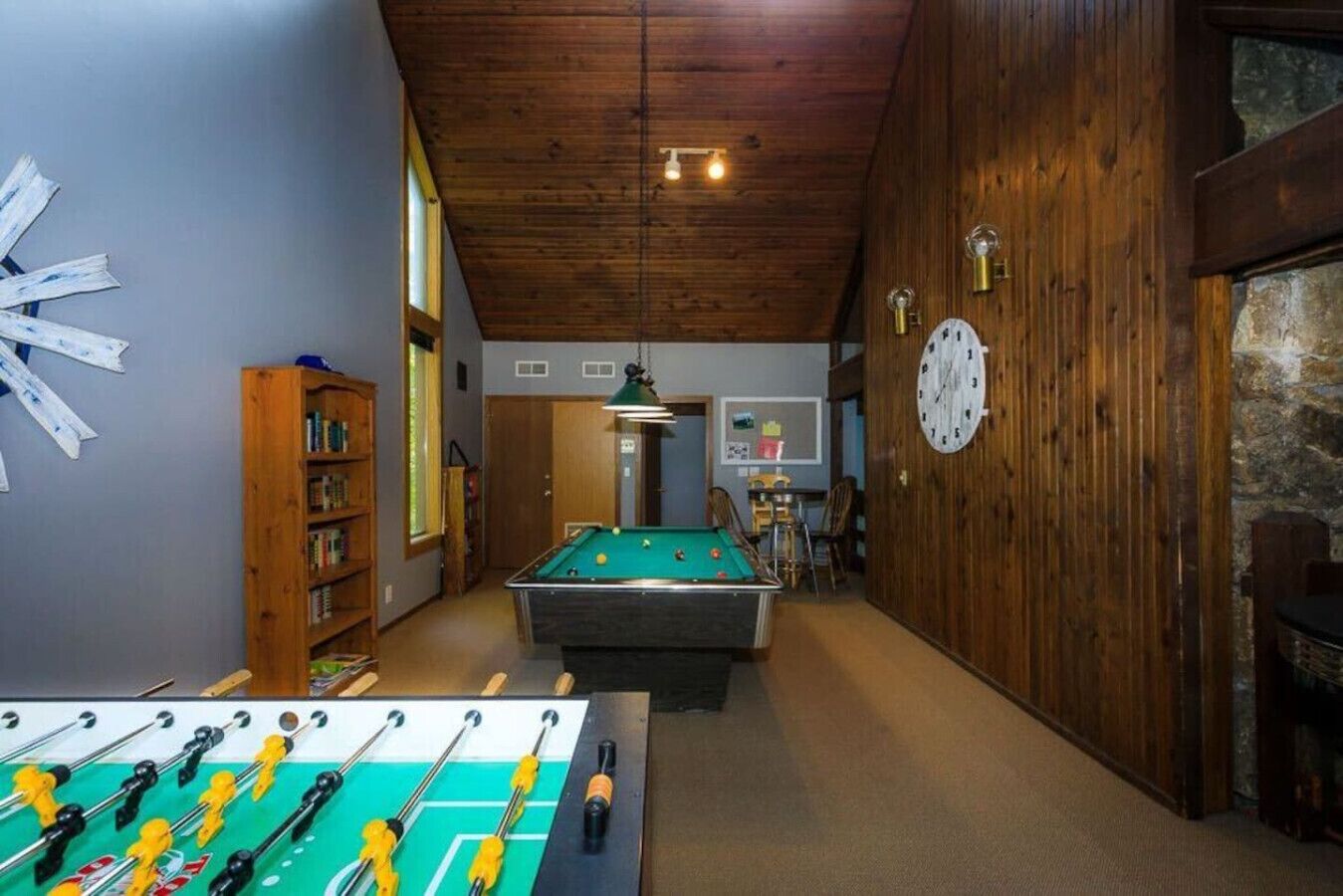 A room with a pool table, foosball table, wooden bookshelves, a clock, and a stone wall.