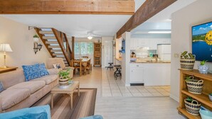 A spacious living area with a high ceiling, wooden beams, a sofa, a coffee table, and a kitchen area with white cabinets and a large window.
