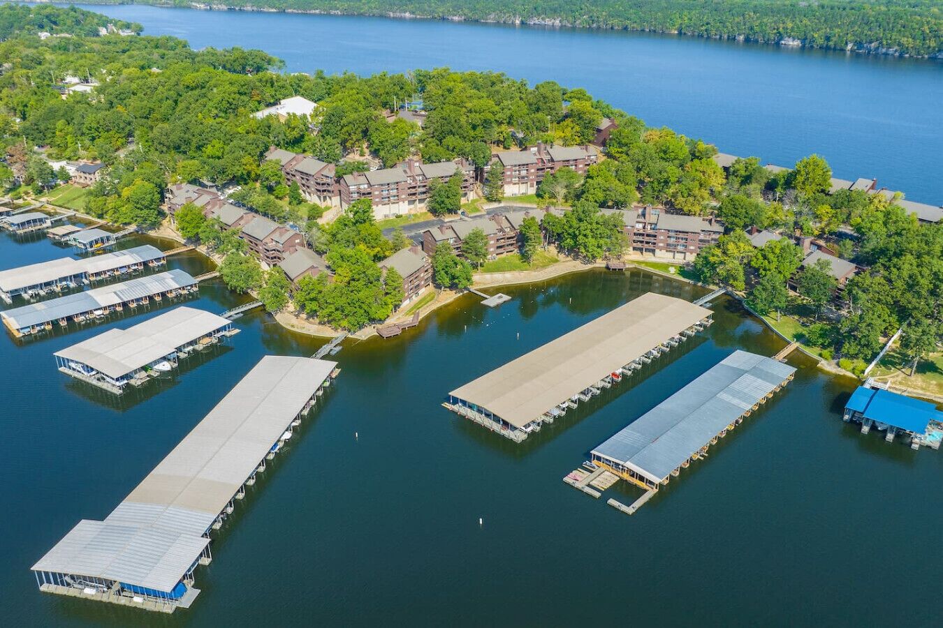 An aerial view of a lakeside property with multiple docked boats, a marina, and residential buildings surrounded by lush greenery.