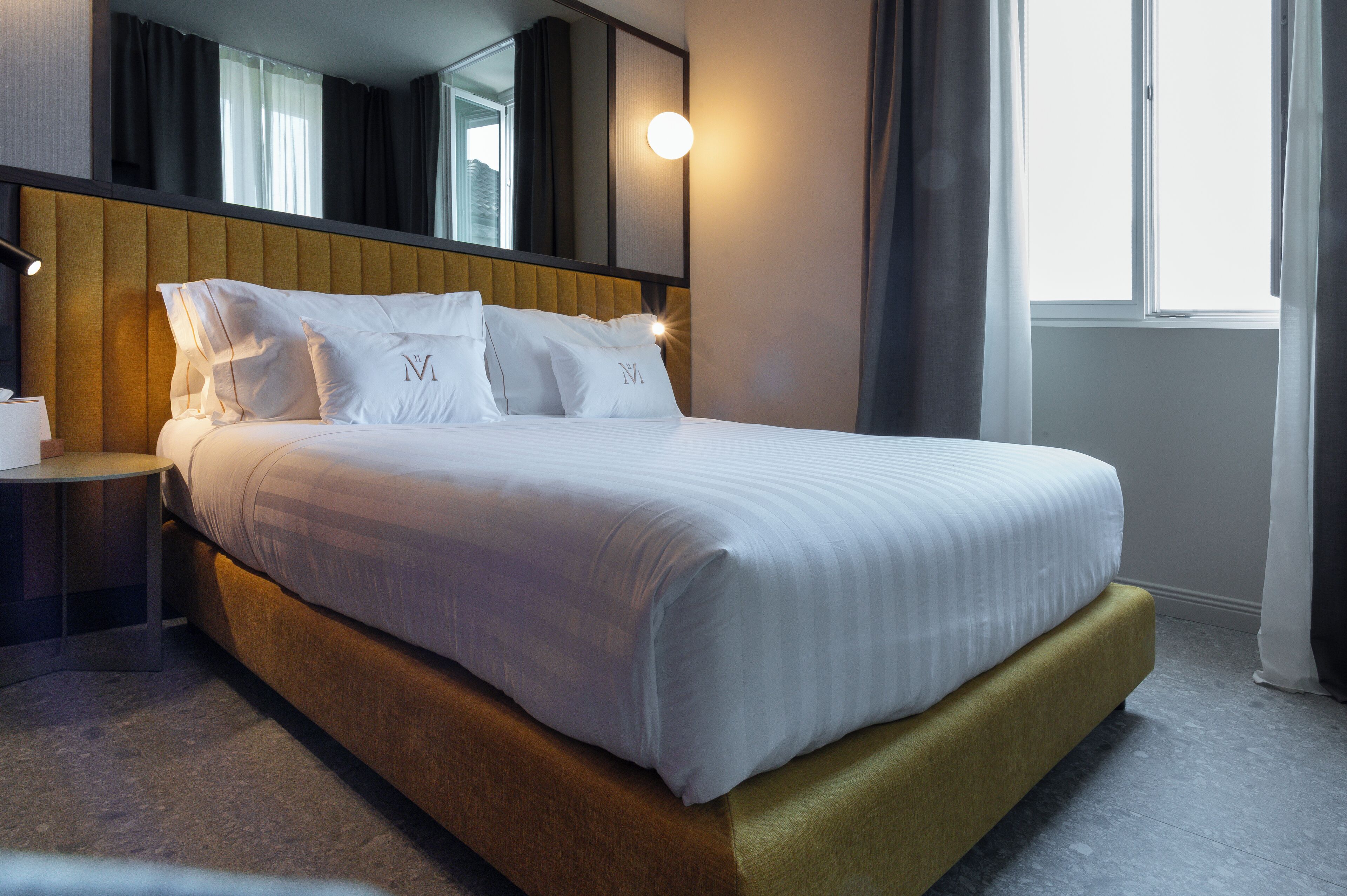 A neatly made bed with white linens and pillows, a wooden headboard, a bedside table with a lamp, and a large window with curtains.