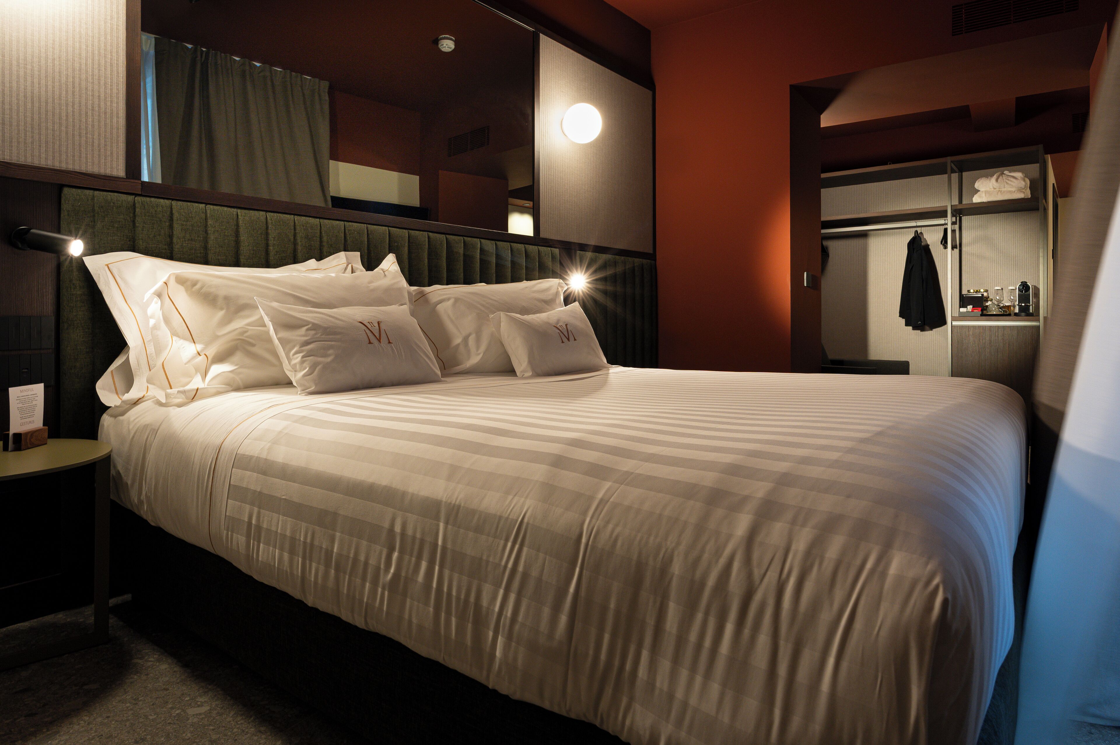 Una cama bien hecha con sábanas y almohadones blancos, una mesita de noche con lámpara, y un espejo sobre la cama.