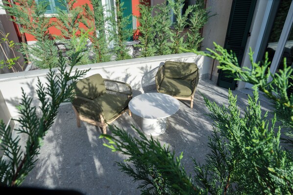 Un patio con dos sillas de mimbre y una mesa redonda, rodeado de plantas verdes.