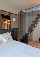 A modern hotel room with a large bed, a mirror, a small table with a book, and a staircase with a metal railing.
